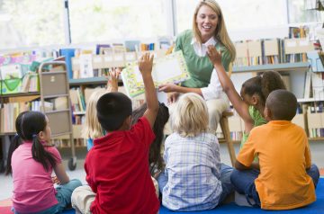 A classroom with 4 small children sitting down with their hands up and a teacher choosing which student to call on